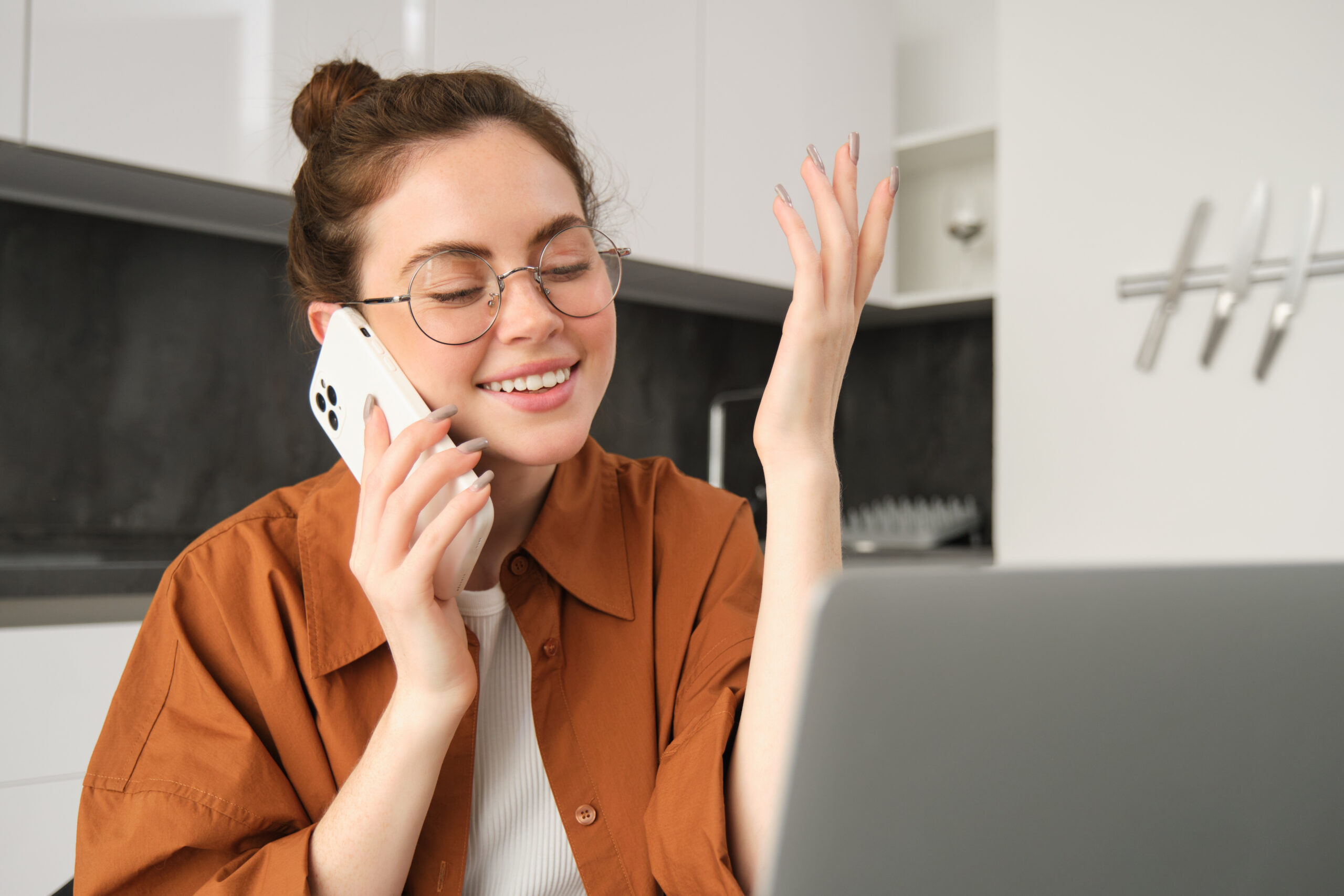 Portrait of young businesswoman, self-employed lady working from home, making phone calls to clients and smiling, using laptop in kitchen